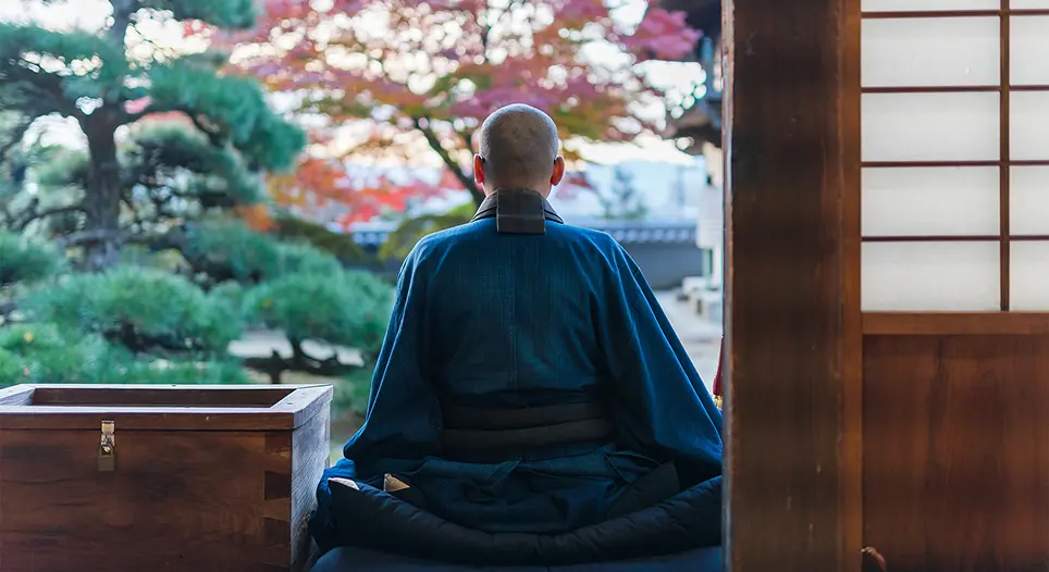 Man in traditional Japanese attire sitting cross-legged facing a garden in a tatami room.