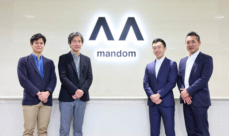 企業ロゴの前に立つ株式会社Mandomの社員