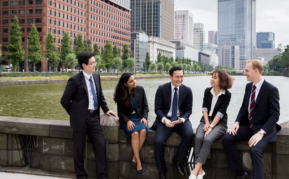 Five business professionals from diverse backgrounds sitting and conversing along a riverside in an urban cityscape