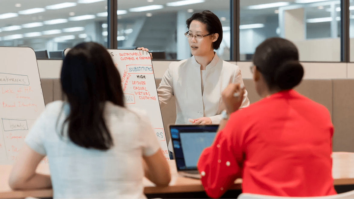 Business professionals engaged in a collaborative discussion around a table, using their laptop PCs with a whiteboard in the background