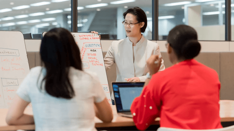 Business professionals engaged in a collaborative discussion around a table, using their laptop PCs with a whiteboard in the background