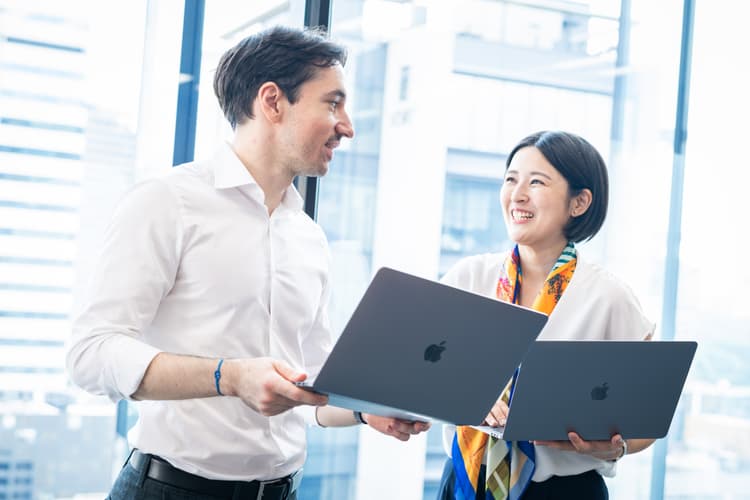 Two business professionals, a man and a woman, are smiling and having a conversation while holding laptops in a modern office with large glass windows in the background.