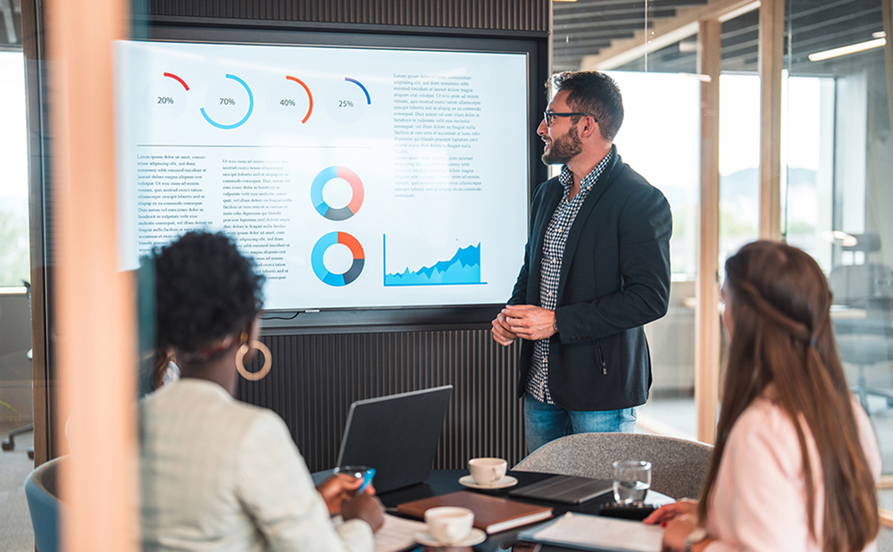 Business professional presenting charts and data on a screen to a group in a meeting room setting
