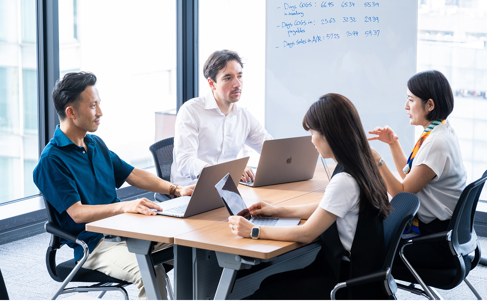 Four business professionals engaged in a collaborative discussion around a table, using their laptop PCs with a whiteboard in the background